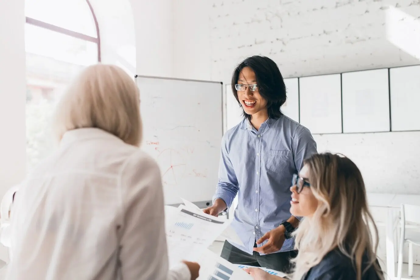 Портрет со спины: блондинка в стильной рубашке читает документы, а азиатский мужчина что‑то ей говорит.